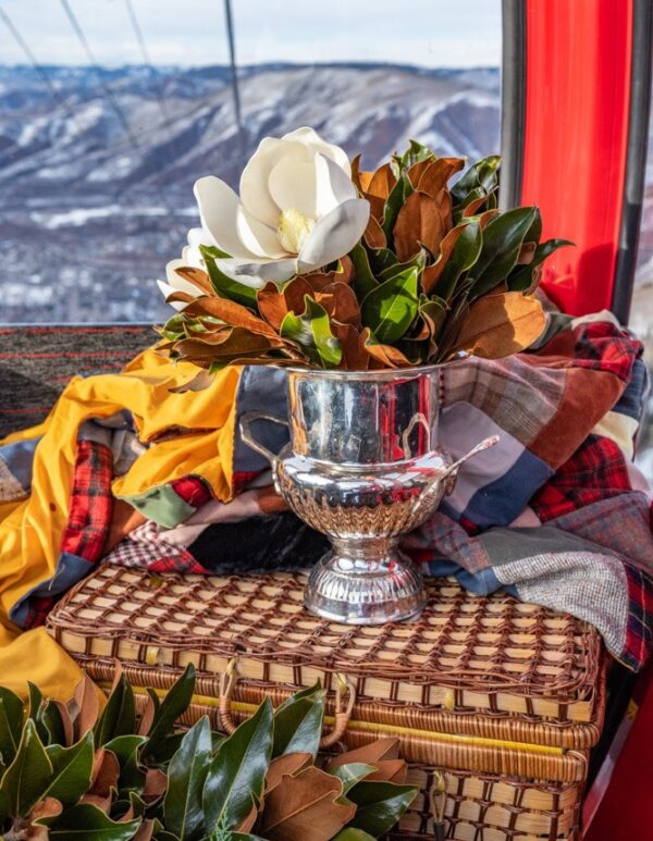 Bright white magnolia flowers in a silver vase on a woven basket, with colorful fabrics and mountain.