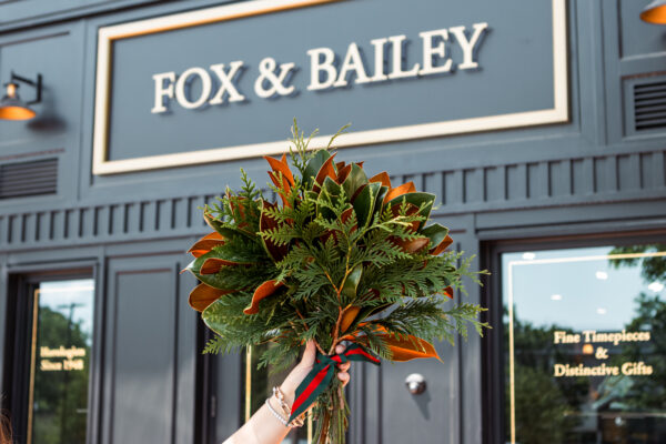 A lush bouquet of mixed greenery and orange flowers held in front of Fox & Bailey storefront.