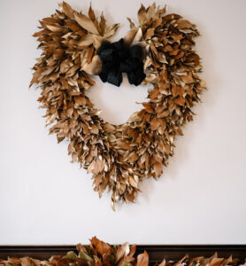 Brown leaf heart-shaped wreath with black bow hanging on a white wall.