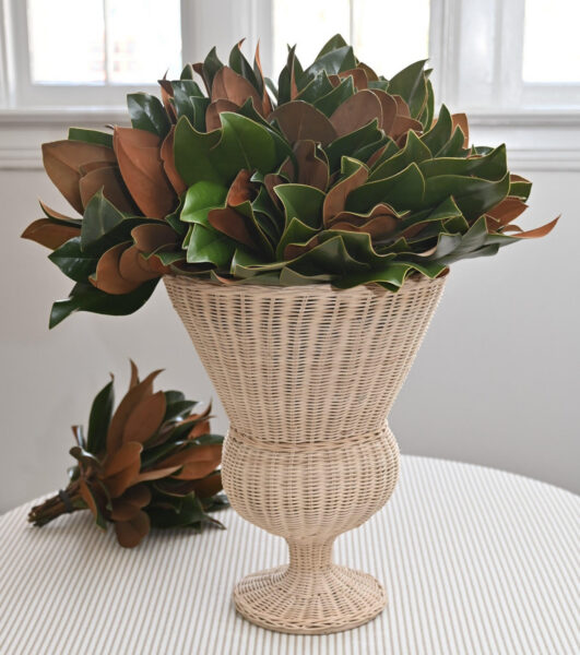 Magnolia leaves in a woven basket vase on a white table with a bright window background.